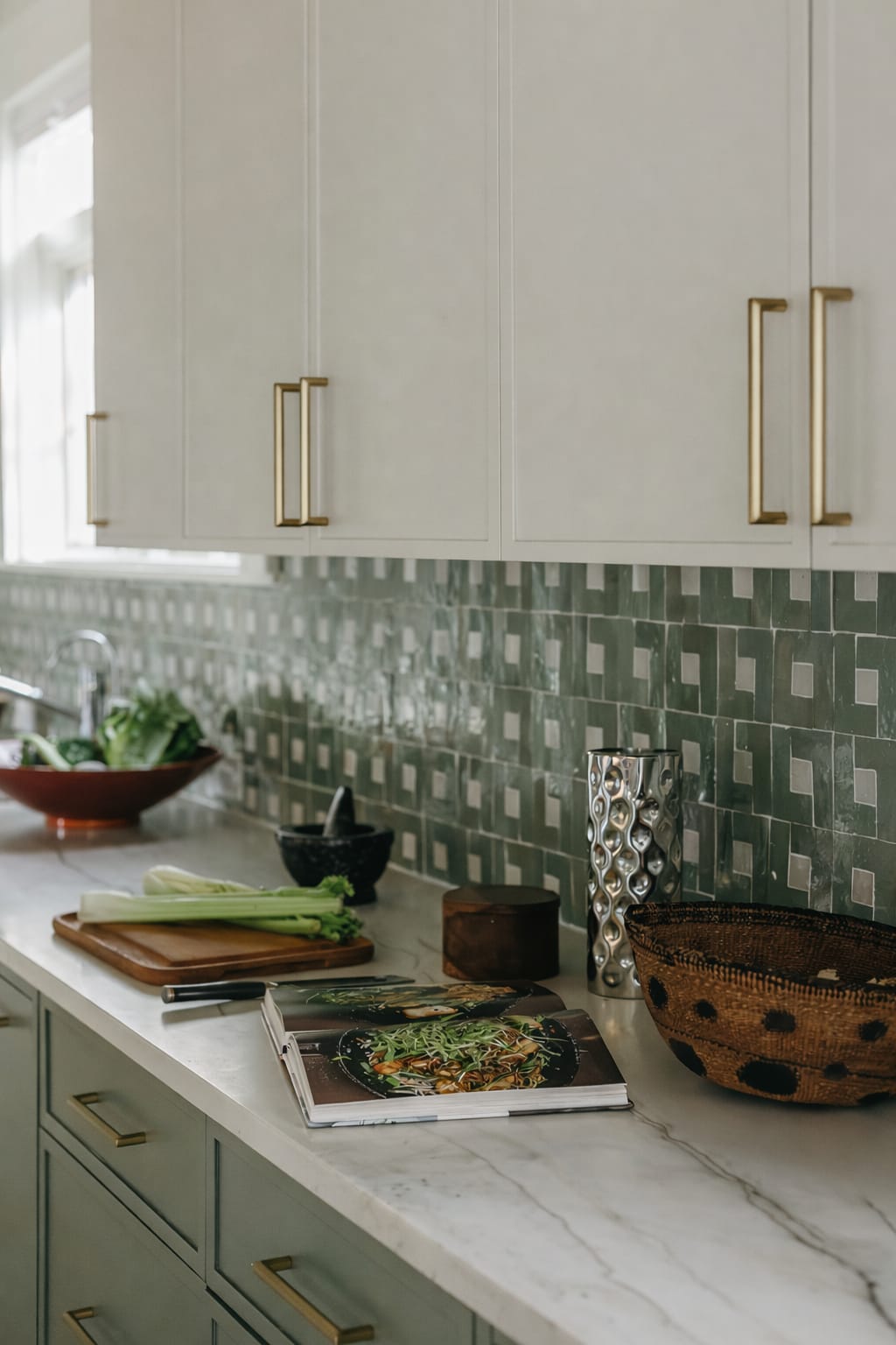 Close view of a green stacked-tile backsplash with brass cabinet hardware, a wooden board and fresh vegetables