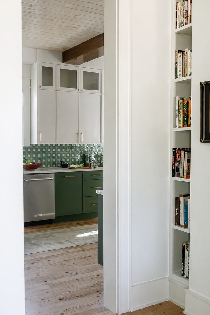 View through a doorway into a green kitchen with glass-front upper cabinets, stainless dishwasher and built-in bookshelves