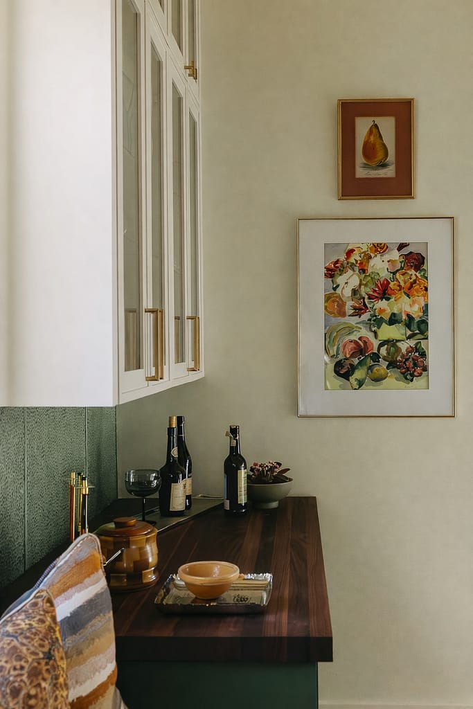 Green tiled wainscot with framed floral artwork above a console table set with wine bottles