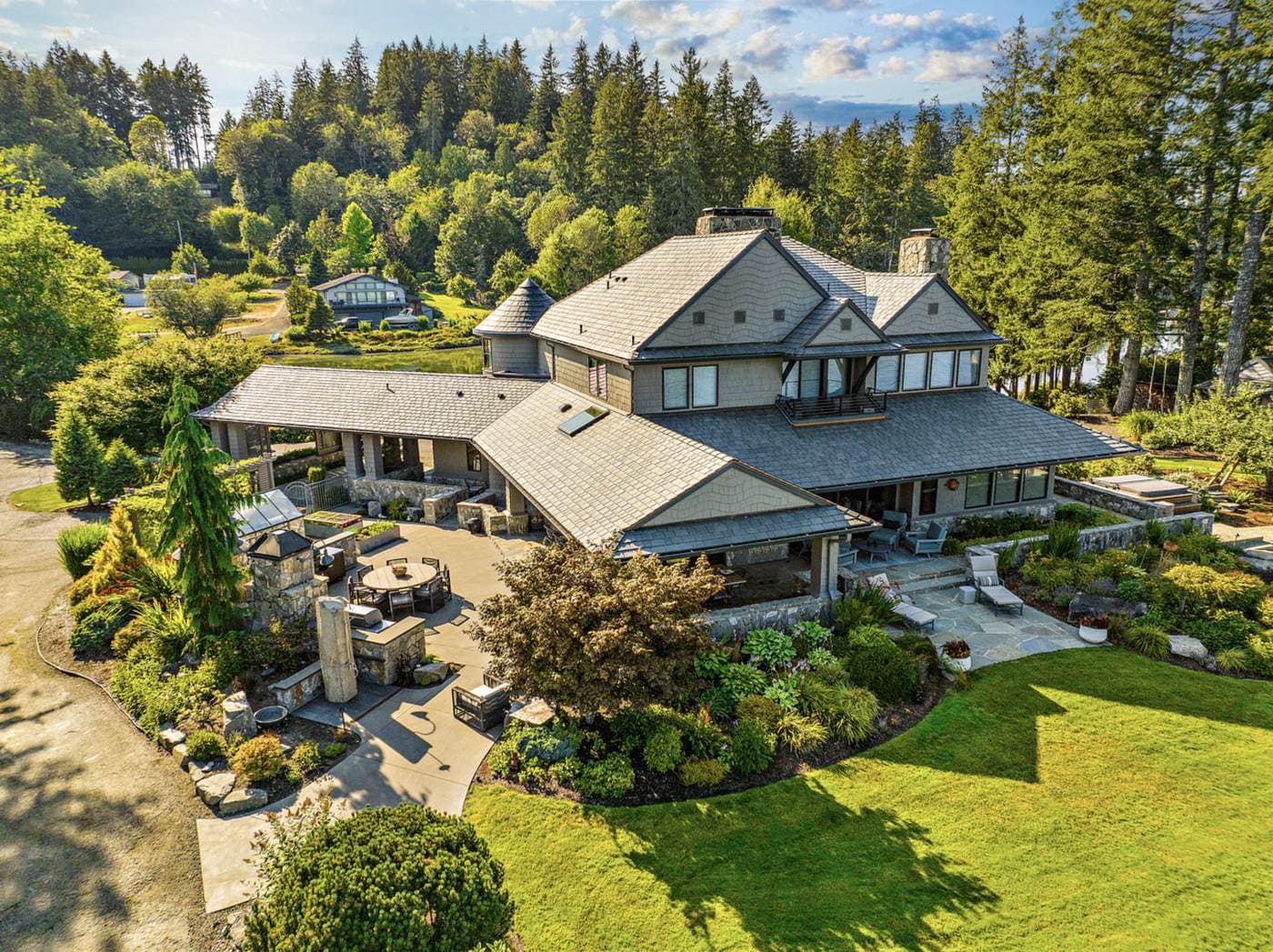 Aerial view of a custom shingle-style home with a stone patio, outdoor seating and landscaped grounds among evergreen trees